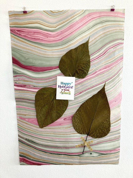 a white greeting card with rainbow foil serif text reading "Happy HoliGays! to You and Yours" is positioned slightly askew and off-center on a large piece of marbled paper with maroon, cream, and green streaks. Three large, dried catalpa leaves surround the card. The stem of one leaf is partially covered with an "x" of golden washi tape.